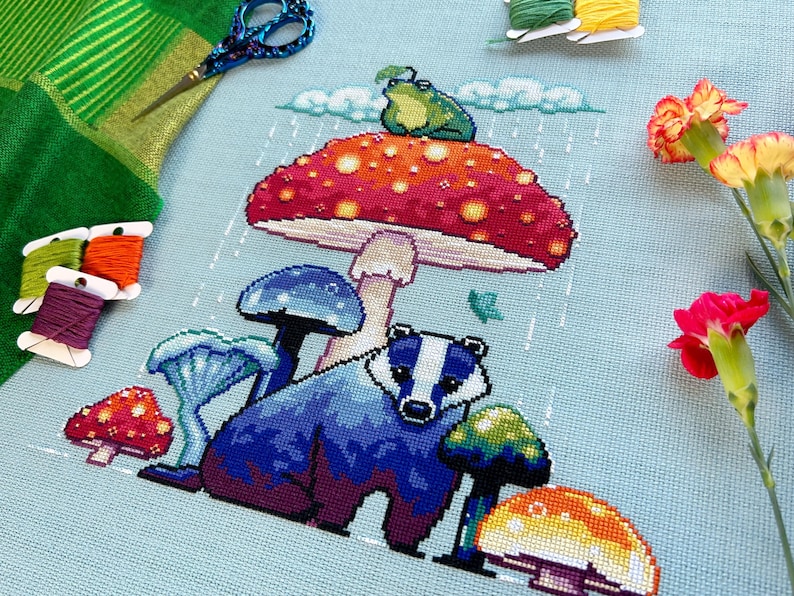 May include: A cross-stitch embroidery featuring a badger standing under a red and yellow mushroom with a green frog on top. The mushroom is surrounded by other mushrooms in shades of blue, green, and yellow. The background is a light blue canvas with white raindrops.