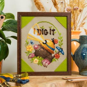 Peut inclure: Une broderie au point de croix encadr&eacute;e repr&eacute;sentant une taupe brune portant un casque de chantier jaune, tenant une pelle bleue, entour&eacute;e d'une couronne de feuilles vertes et de fleurs. Le texte "I DIG IT" est brod&eacute; en brun au-dessus.