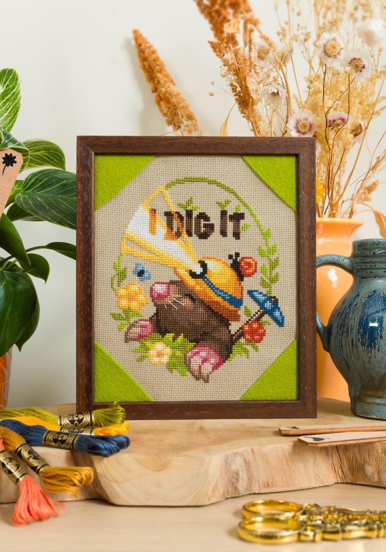 Peut inclure: Une broderie au point de croix encadr&eacute;e repr&eacute;sentant une taupe brune portant un casque de chantier jaune, tenant une pelle bleue, entour&eacute;e d'une couronne de feuilles vertes et de fleurs. Le texte "I DIG IT" est brod&eacute; en brun au-dessus.