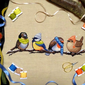 Flatlay of Winter Birds cross stitch pattern. Stitched item is surrounded by decorations. Finished piece is of medium to large size. Colors are yellow, brown, orange, blue, green, white. There are four birds sitting on a branch wearing winter gear.