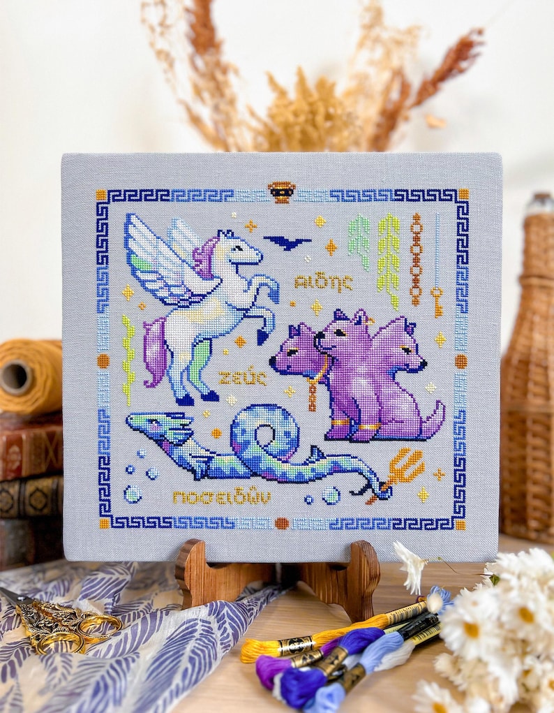 May include: A cross-stitch embroidery featuring mythological creatures. The design includes a winged unicorn, a three-headed dog, and a sea serpent, all in vibrant colors. The artwork is framed with a Greek key border and includes Greek text.