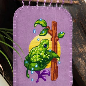 Peut inclure: Une tenture murale en tissu violet avec une broderie au point de croix repr&eacute;sentant une grenouille verte sur une branche brune devant un cercle jaune. La grenouille est entour&eacute;e de feuilles vertes et de petites gouttes d'eau bleues.
