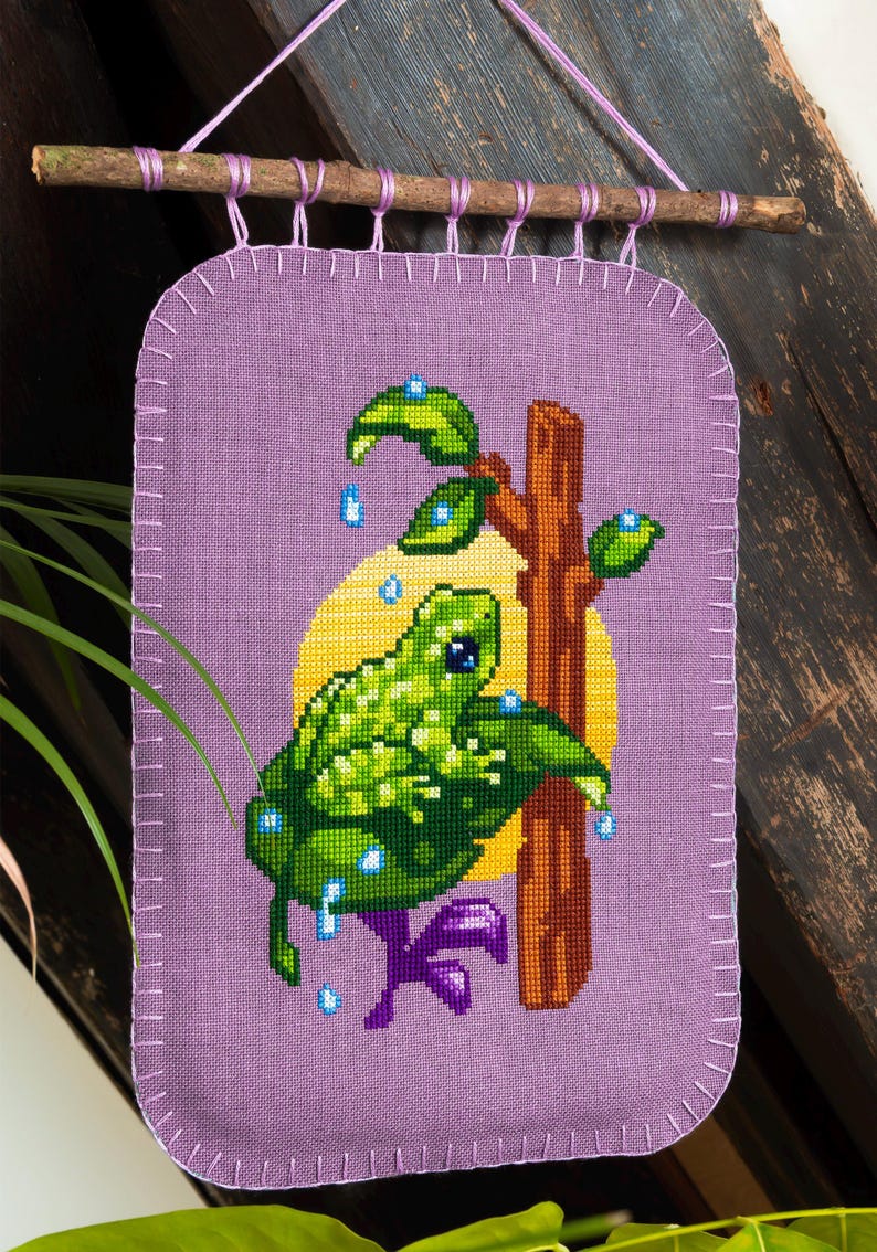 Peut inclure: Une tenture murale en tissu violet avec une broderie au point de croix repr&eacute;sentant une grenouille verte sur une branche brune devant un cercle jaune. La grenouille est entour&eacute;e de feuilles vertes et de petites gouttes d'eau bleues.