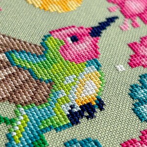 May include: Close-up of a cross-stitch design featuring a hummingbird. The bird has a pink head, green body, and blue wings. The background is a light green fabric, with other colorful stitched elements.