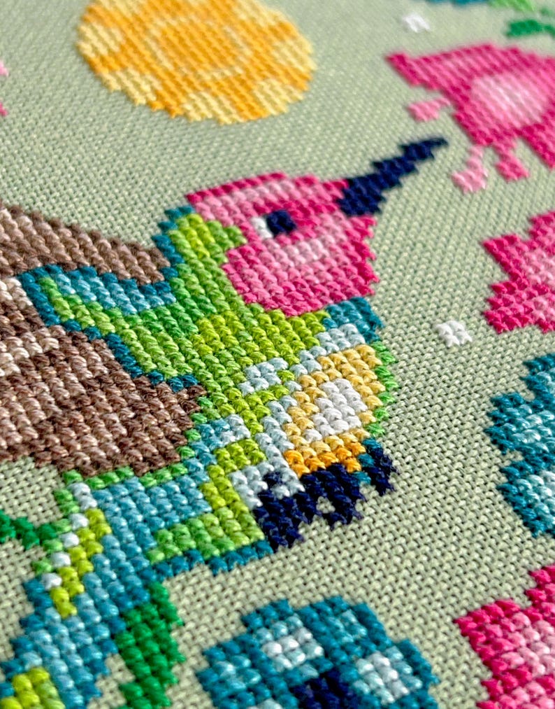May include: Close-up of a cross-stitch design featuring a hummingbird. The bird has a pink head, green body, and blue wings. The background is a light green fabric, with other colorful stitched elements.