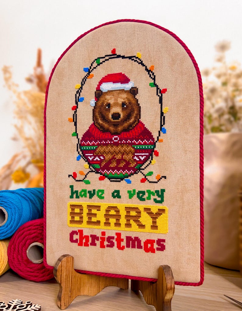 May include: A cross-stitch design featuring a brown bear wearing a Santa hat and a red sweater, surrounded by Christmas lights. The text "have a very BEARY Christmas" is stitched below the bear. The design is framed in red and displayed on a wooden stand.
