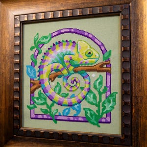 May include: Cross-stitch artwork of a chameleon in a wooden frame. The chameleon is depicted in shades of yellow, purple, and blue, perched on a branch. Green leaves and a purple border surround the reptile, set against a light green background.