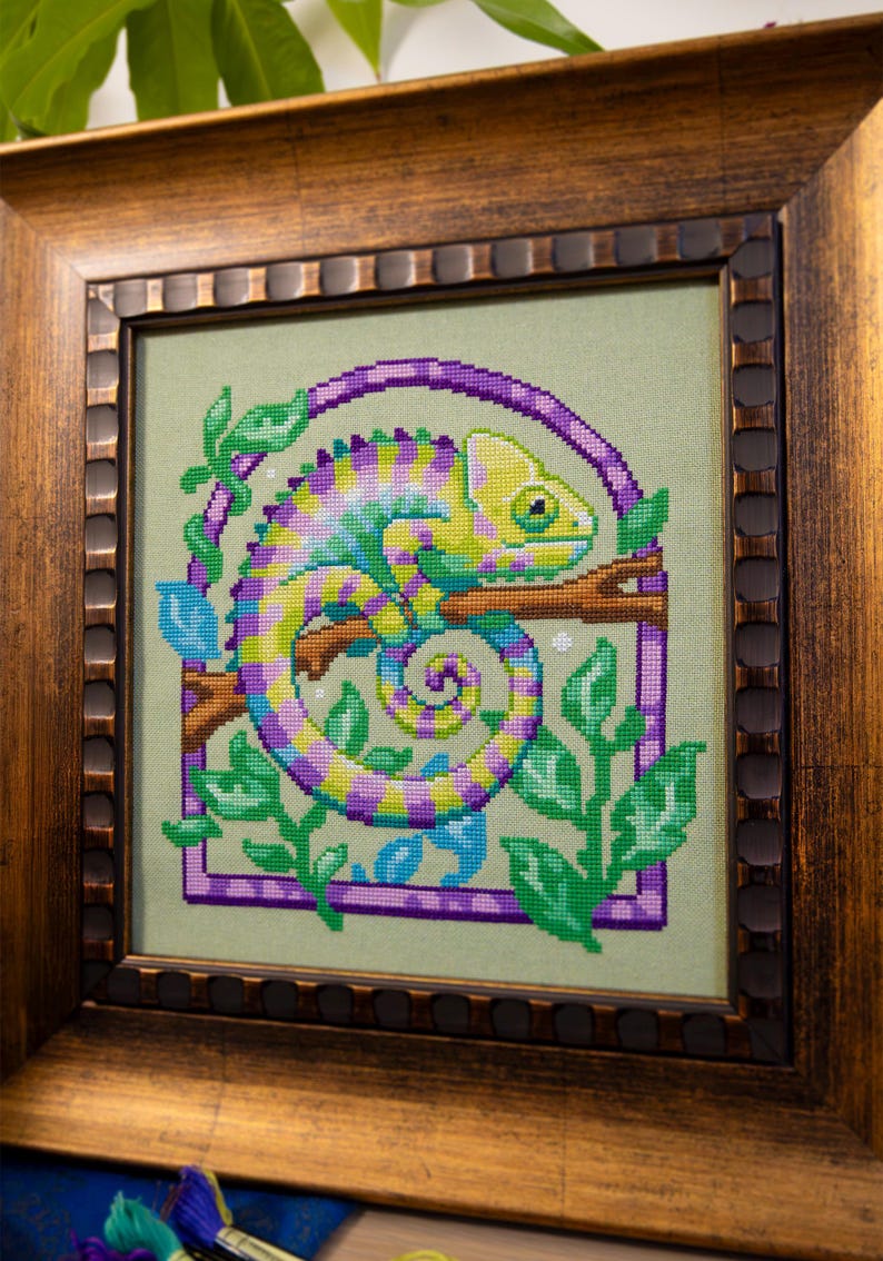 May include: Cross-stitch artwork of a chameleon in a wooden frame. The chameleon is depicted in shades of yellow, purple, and blue, perched on a branch. Green leaves and a purple border surround the reptile, set against a light green background.