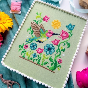 May include: A cross-stitch piece featuring a hummingbird in flight, surrounded by colorful flowers and a yellow sun. The artwork is framed with a light green fabric and a white pom-pom trim. The color palette includes pink, blue, green, and brown.