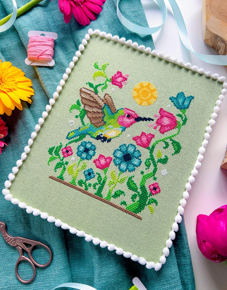 May include: A cross-stitch piece featuring a hummingbird in flight, surrounded by colorful flowers and a yellow sun. The artwork is framed with a light green fabric and a white pom-pom trim. The color palette includes pink, blue, green, and brown.