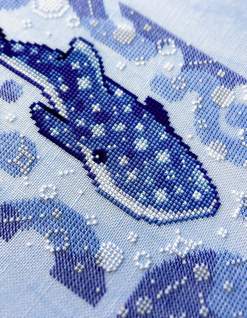 May include: A cross-stitch design of a whale shark in shades of blue and white. The whale shark is the focal point, with a detailed pattern of stitches. The background features abstract shapes and bubbles, all in a light blue hue.