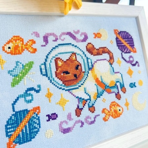 May include: Cross-stitch art of a cat in a spacesuit, surrounded by space-themed elements like planets, fish, and stars. The artwork is framed in a white wooden frame, with a light blue background. The cat is brown and white.