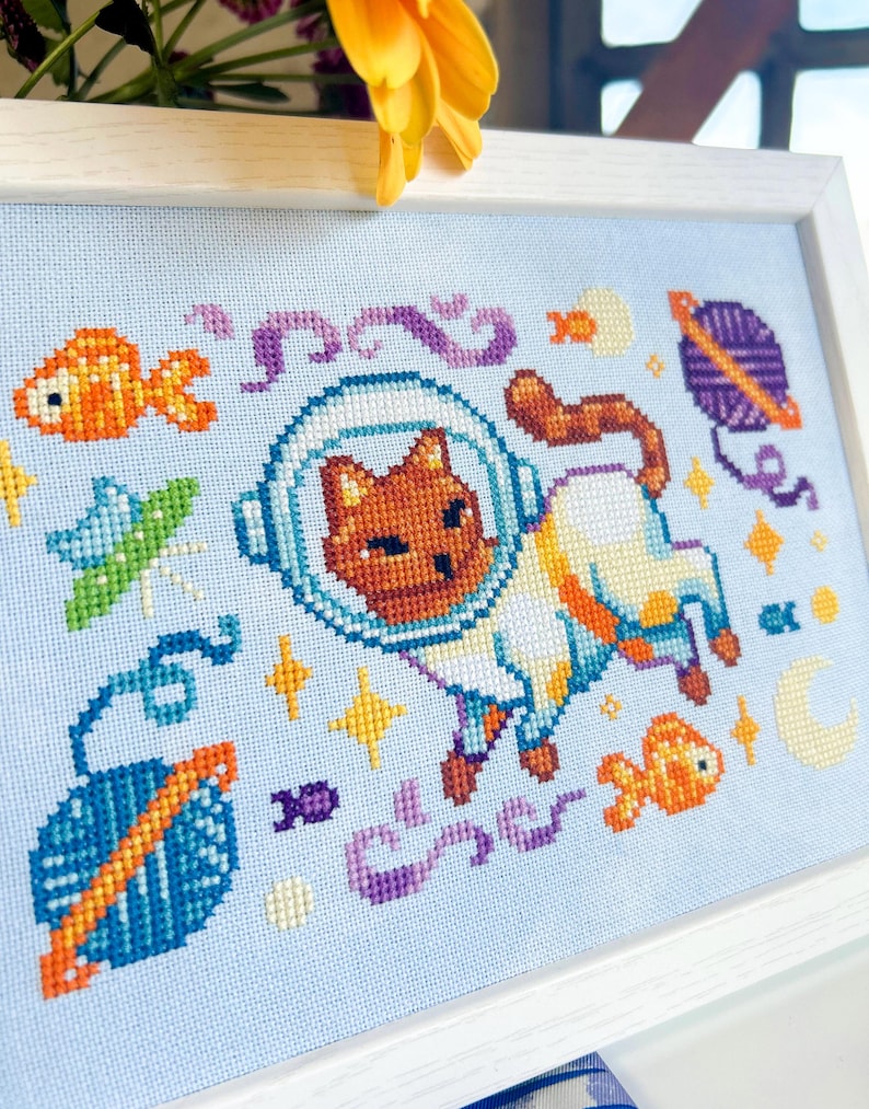 May include: Cross-stitch art of a cat in a spacesuit, surrounded by space-themed elements like planets, fish, and stars. The artwork is framed in a white wooden frame, with a light blue background. The cat is brown and white.