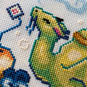 May include: A cross-stitch embroidery on light blue Aida fabric featuring a green creature with blue accents, a small kite, and orange and blue shapes. The design uses various colored threads to create a textured, pixelated appearance on the grid.