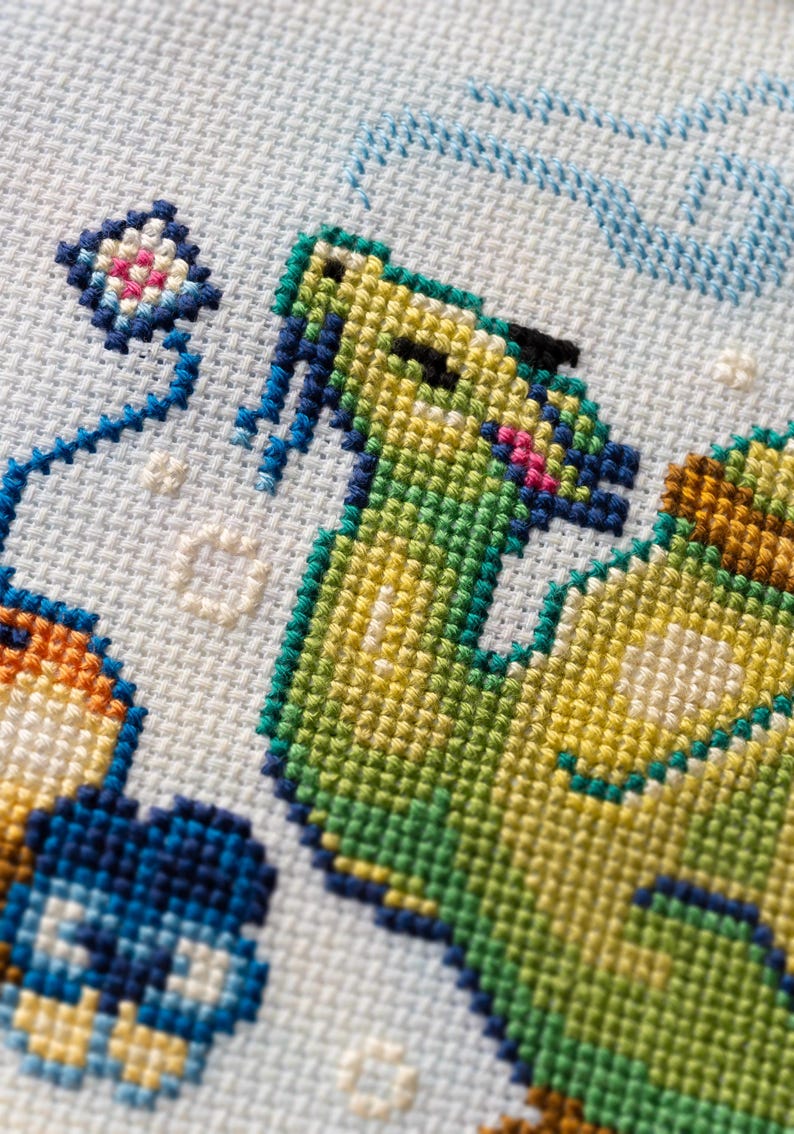 May include: A cross-stitch embroidery on light blue Aida fabric featuring a green creature with blue accents, a small kite, and orange and blue shapes. The design uses various colored threads to create a textured, pixelated appearance on the grid.
