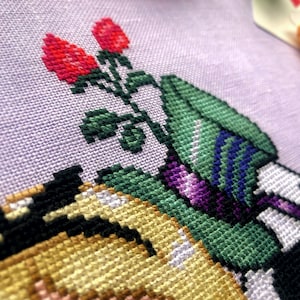 Closeup of top right side of cross stitch pattern. Here the hat of the Mad Hatter is visible. It is tall and green with blue markings, and with a purple ribbon wrapped around it. Behind it are more red roses.