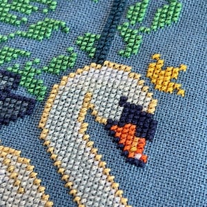 Puede incluir: Primer plano de un bordado de punto de cruz con dos cisnes blancos con detalles amarillos y naranjas. Los cisnes est&aacute;n rodeados de hojas verdes sobre un fondo azul.