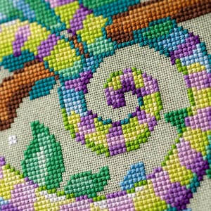 May include: Close-up of a cross-stitch design featuring a colorful chameleon. The chameleon is depicted in shades of green, purple, yellow, and blue, with a brown branch. The cross-stitch is on a light beige fabric.