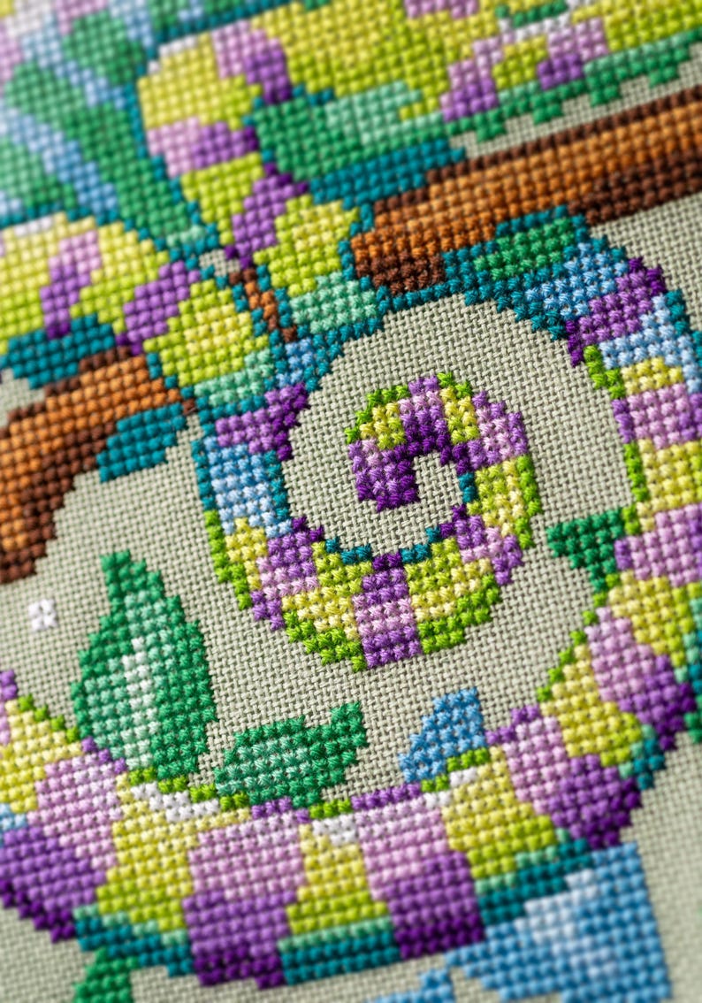 May include: Close-up of a cross-stitch design featuring a colorful chameleon. The chameleon is depicted in shades of green, purple, yellow, and blue, with a brown branch. The cross-stitch is on a light beige fabric.