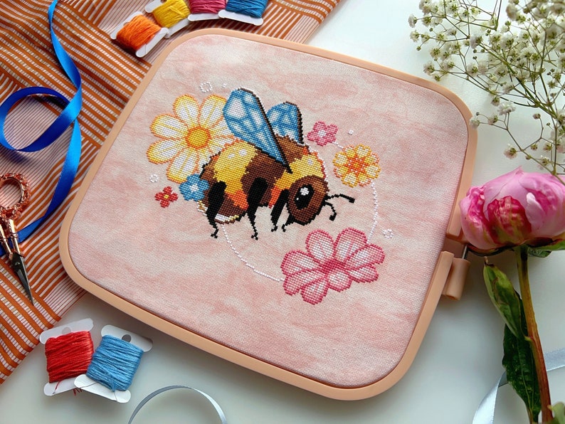 May include: A pink and white embroidery hoop with a cross-stitch design of a bee surrounded by flowers. The bee is yellow and black with blue wings. The flowers are pink, white, and yellow.