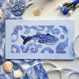 May include: Cross-stitch art featuring a whale shark in shades of blue, surrounded by stylized waves and bubbles. The artwork is on light blue fabric, with a patterned blue and white fabric, seashells, scissors, and embroidery floss in the background.