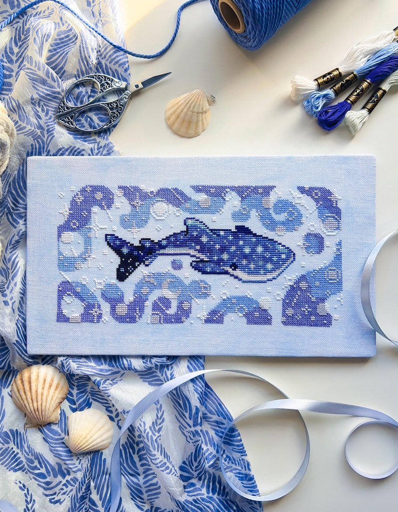 May include: Cross-stitch art featuring a whale shark in shades of blue, surrounded by stylized waves and bubbles. The artwork is on light blue fabric, with a patterned blue and white fabric, seashells, scissors, and embroidery floss in the background.