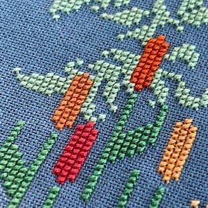Puede incluir: Primer plano de una tela azul con un dise&ntilde;o de punto de cruz con hilos verdes, naranjas y rojos. El dise&ntilde;o es abstracto y se asemeja a una planta o flor estilizada.