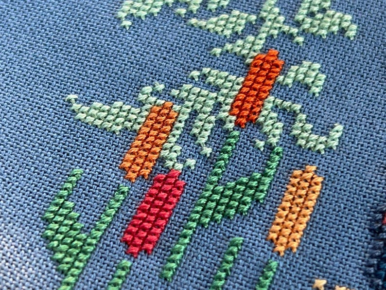 Puede incluir: Primer plano de una tela azul con un dise&ntilde;o de punto de cruz con hilos verdes, naranjas y rojos. El dise&ntilde;o es abstracto y se asemeja a una planta o flor estilizada.