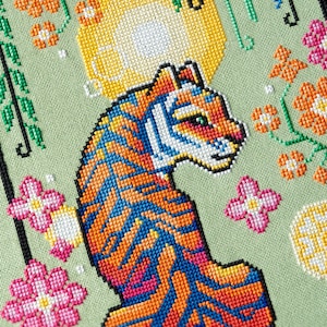 May include: A colorful cross-stitch design featuring a tiger with orange, blue, and pink stripes. The artwork includes floral accents in pink and orange, set against a light green background. The tiger is the central focus of the piece.