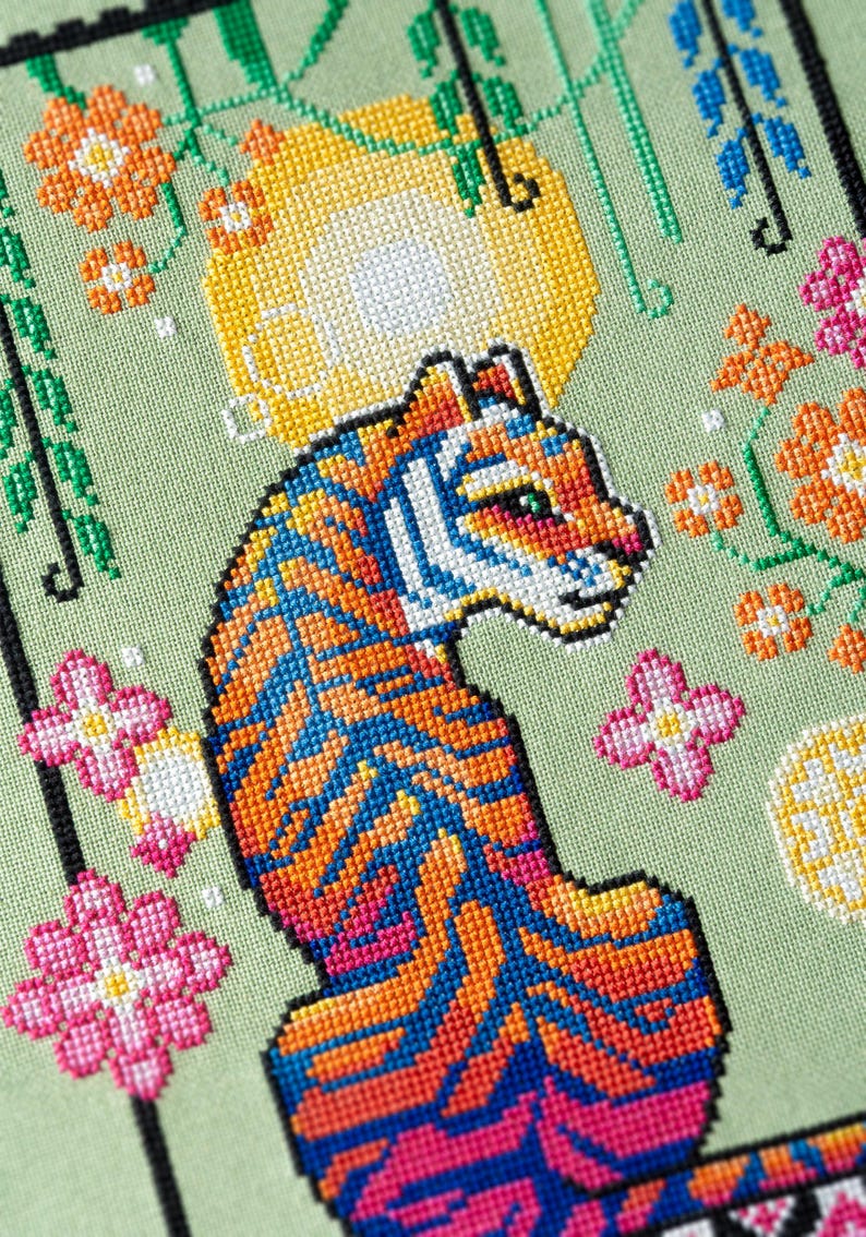 May include: A colorful cross-stitch design featuring a tiger with orange, blue, and pink stripes. The artwork includes floral accents in pink and orange, set against a light green background. The tiger is the central focus of the piece.