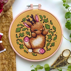 May include: A cross-stitch embroidery hoop with a yellow background and a brown bunny with antlers sleeping surrounded by green leaves.