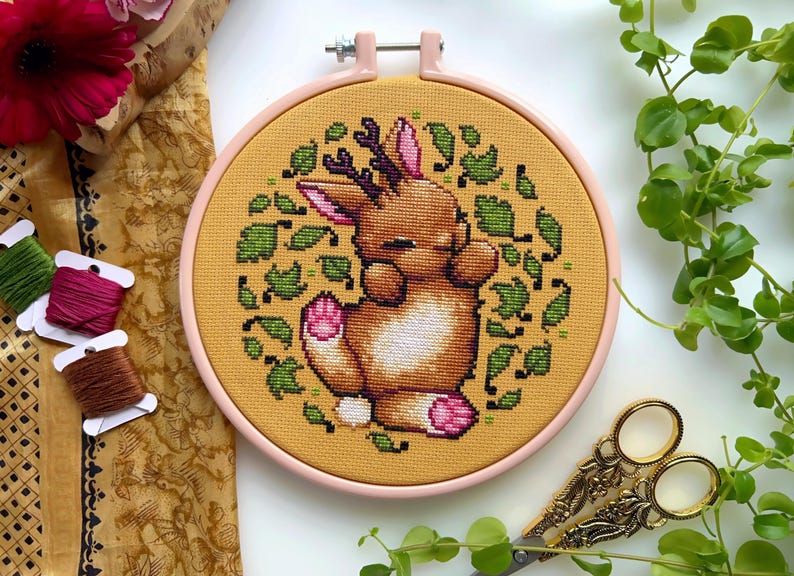 May include: A cross-stitch embroidery hoop with a yellow background and a brown bunny with antlers sleeping surrounded by green leaves.