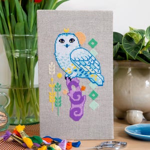 Op de afbeelding: Kruissteekkunstwerk van een sneeuwuil op een paarse tak. De uil is wit en blauw met een gele kroon. De achtergrond is een lichtgrijze stof. Borduurgaren en schaar op de voorgrond.