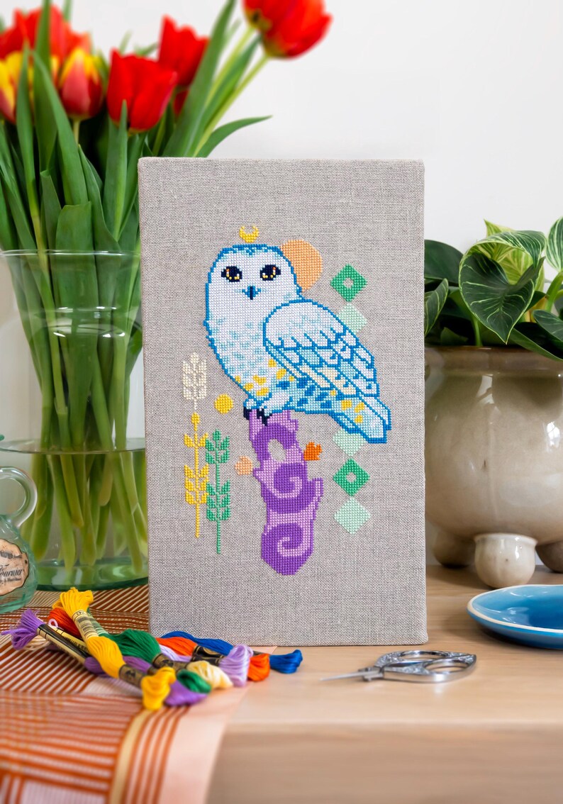 Op de afbeelding: Kruissteekkunstwerk van een sneeuwuil op een paarse tak. De uil is wit en blauw met een gele kroon. De achtergrond is een lichtgrijze stof. Borduurgaren en schaar op de voorgrond.