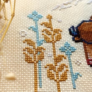 May include: Close-up of a cross-stitch pattern featuring a brown bear with blue glasses, surrounded by gold and blue floral designs.