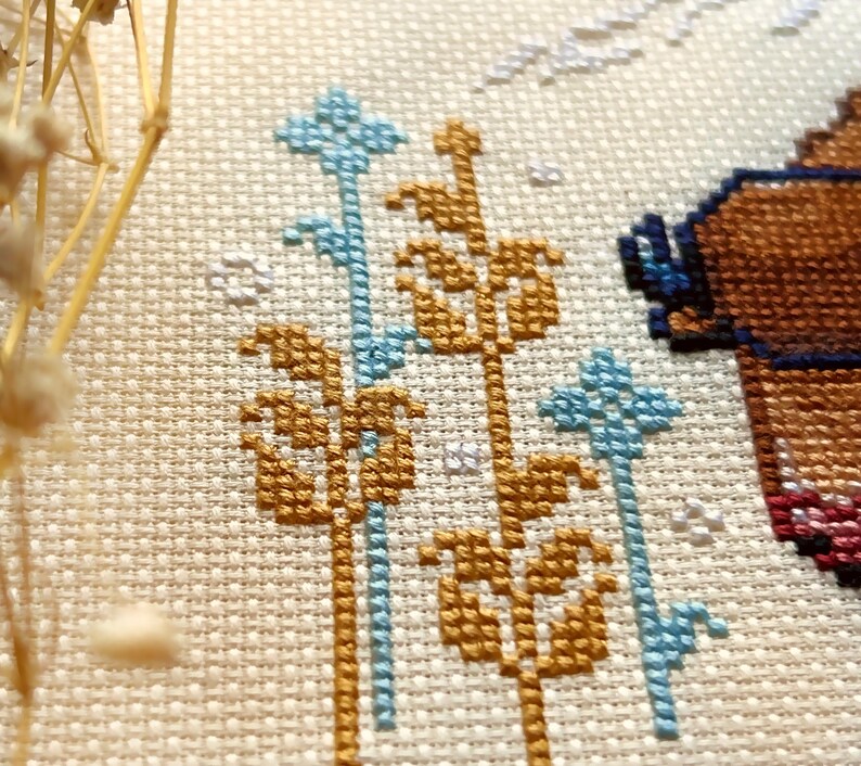 May include: Close-up of a cross-stitch pattern featuring a brown bear with blue glasses, surrounded by gold and blue floral designs.