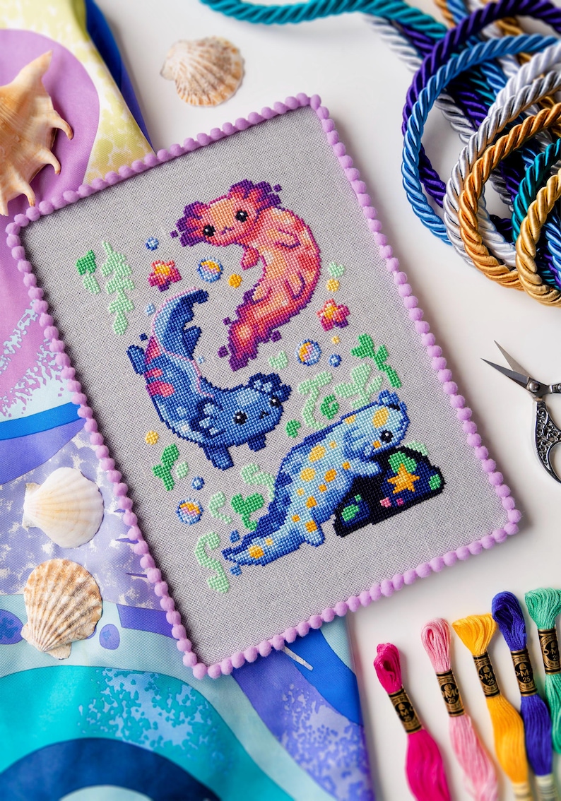May include: Cross-stitch art featuring three colorful axolotls in a whimsical underwater scene. The embroidery is framed with a lavender border and pom-poms. Colorful embroidery floss, seashells, and decorative rope are also visible.