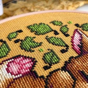 May include: Close-up of a cross-stitch embroidery hoop with a design of a brown and white fox with pink flowers and green leaves on a yellow background.