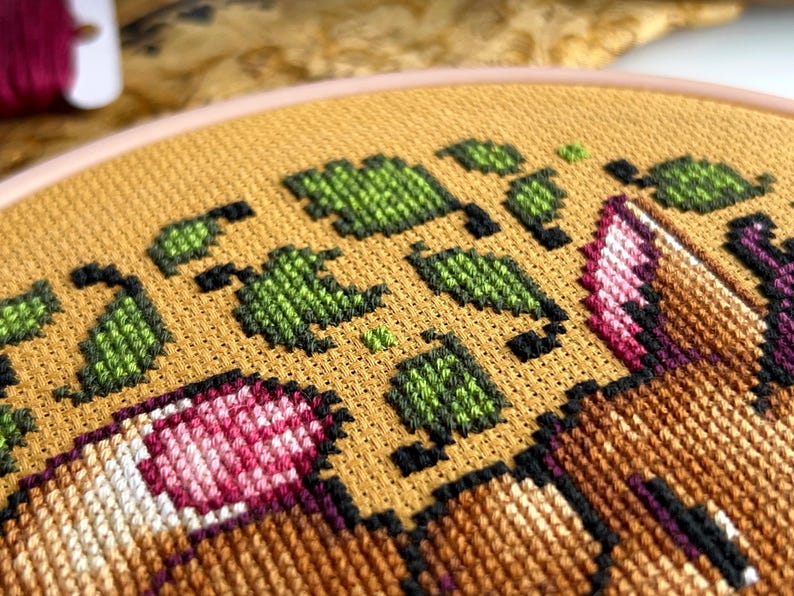 May include: Close-up of a cross-stitch embroidery hoop with a design of a brown and white fox with pink flowers and green leaves on a yellow background.