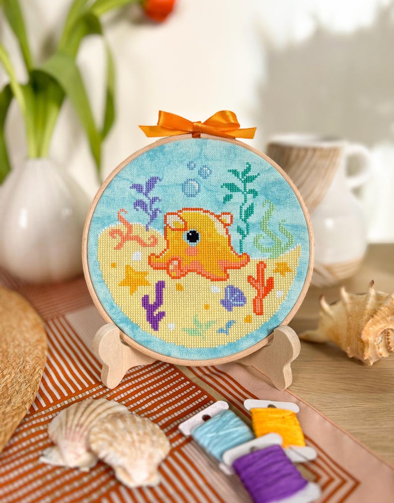 May include: A completed cross-stitch embroidery hoop with a yellow octopus in a blue ocean scene. The hoop is on a wooden stand and there are colorful embroidery floss spools nearby.