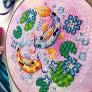 Closeup of Koi Fish cross stitch pattern. Right side is most visible. Finished piece is of medium size. Colors are very bright, and are green, blue, orange, pink and yellow. There are two fish, surrounded by lotus leaves and flowers.