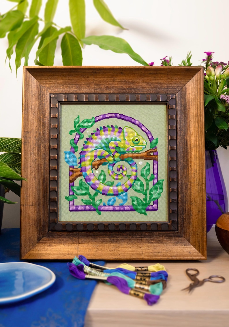 May include: Cross-stitch art of a chameleon in shades of green, yellow, purple, and blue, framed in a wooden frame. The chameleon is perched on a branch, surrounded by green leaves and a purple border. The frame is a warm brown color.