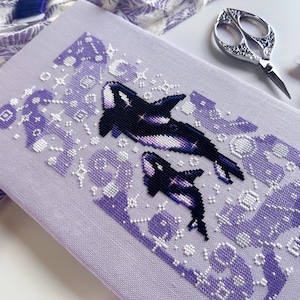 Puede incluir: Un patrón de punto de cruz con dos orcas nadando en un cielo estrellado morado y blanco. Las orcas son negras y blancas con un degradado morado.