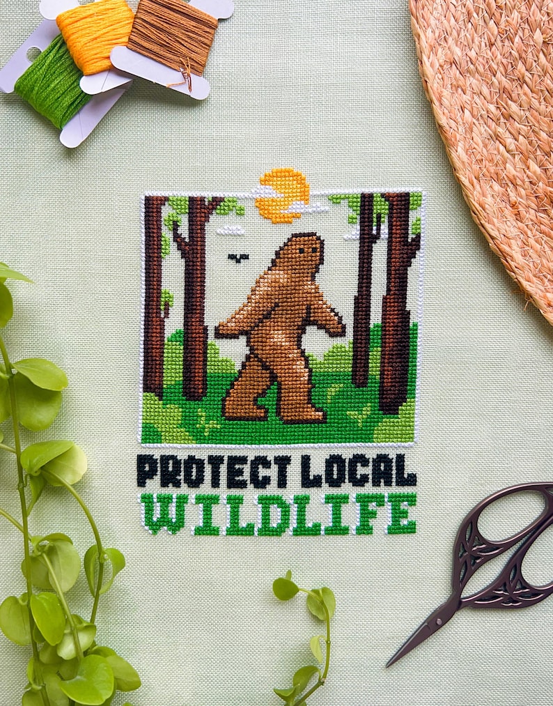 May include: Cross-stitch art featuring a brown Bigfoot walking in a forest scene with a yellow sun. The text "PROTECT LOCAL WILDLIFE" is stitched in green and white. The image is surrounded by green thread, orange thread, brown thread, and a pair of scissors.
