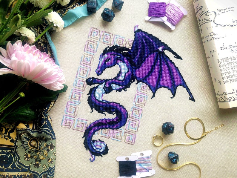 Flat-lay of Dragon cross stitch pattern. Stitched item, surrounded by decorations. Finished piece is of medium to large size. Colors are blue, purple, white, black and pink. Dragon is surrounded by variegated frame. Its head is like that of a horse.