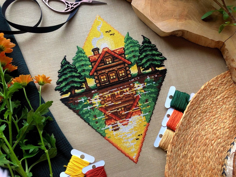 Flat-lay of Lakeside Cabin cross stitch pattern. Stitched item, surrounded by decorations. Finished piece is of large size. Colors are yellow, brown, orange, green, black and white. Scenery contains a sunset, and the cabin is reflected in the water.