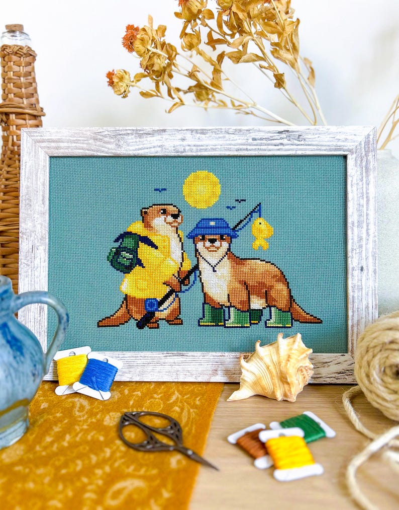 May include: Cross-stitch artwork depicting two otters fishing, set against a teal background with a yellow sun. One otter wears a yellow raincoat and carries a backpack, while the other wears a blue hat and green wellington boots. Framed in a distressed white wooden frame.