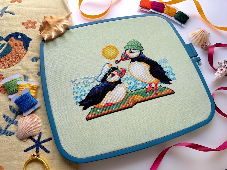 May include: Cross-stitch embroidery of two puffins wearing hats, one with a pipe, on a green background with blue waves and a yellow sun. The puffins are sitting on a book with a star.