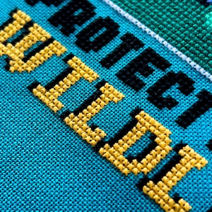 May include: Close-up of a cross-stitch design on turquoise fabric. The words "PROTECT WILD" are stitched in yellow and black thread. The design includes a green and white border.
