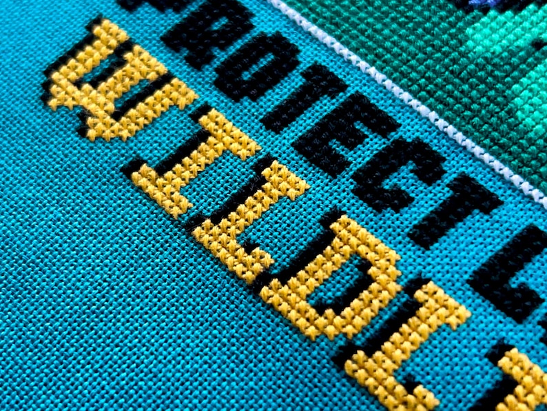 May include: Close-up of a cross-stitch design on turquoise fabric. The words "PROTECT WILD" are stitched in yellow and black thread. The design includes a green and white border.